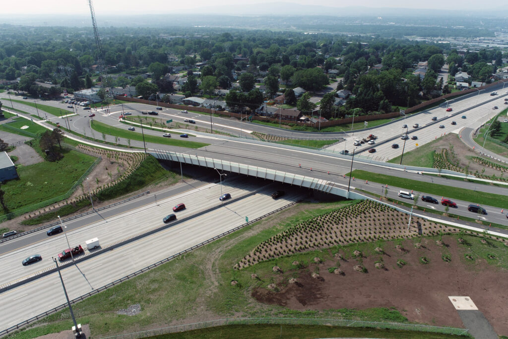 Vue aérienne d'un échangeur autoroutier avec des voitures circulant sur plusieurs voies, un viaduc et des espaces paysagers entourant les routes. Un quartier résidentiel et des collines sont visibles à l'arrière-plan.