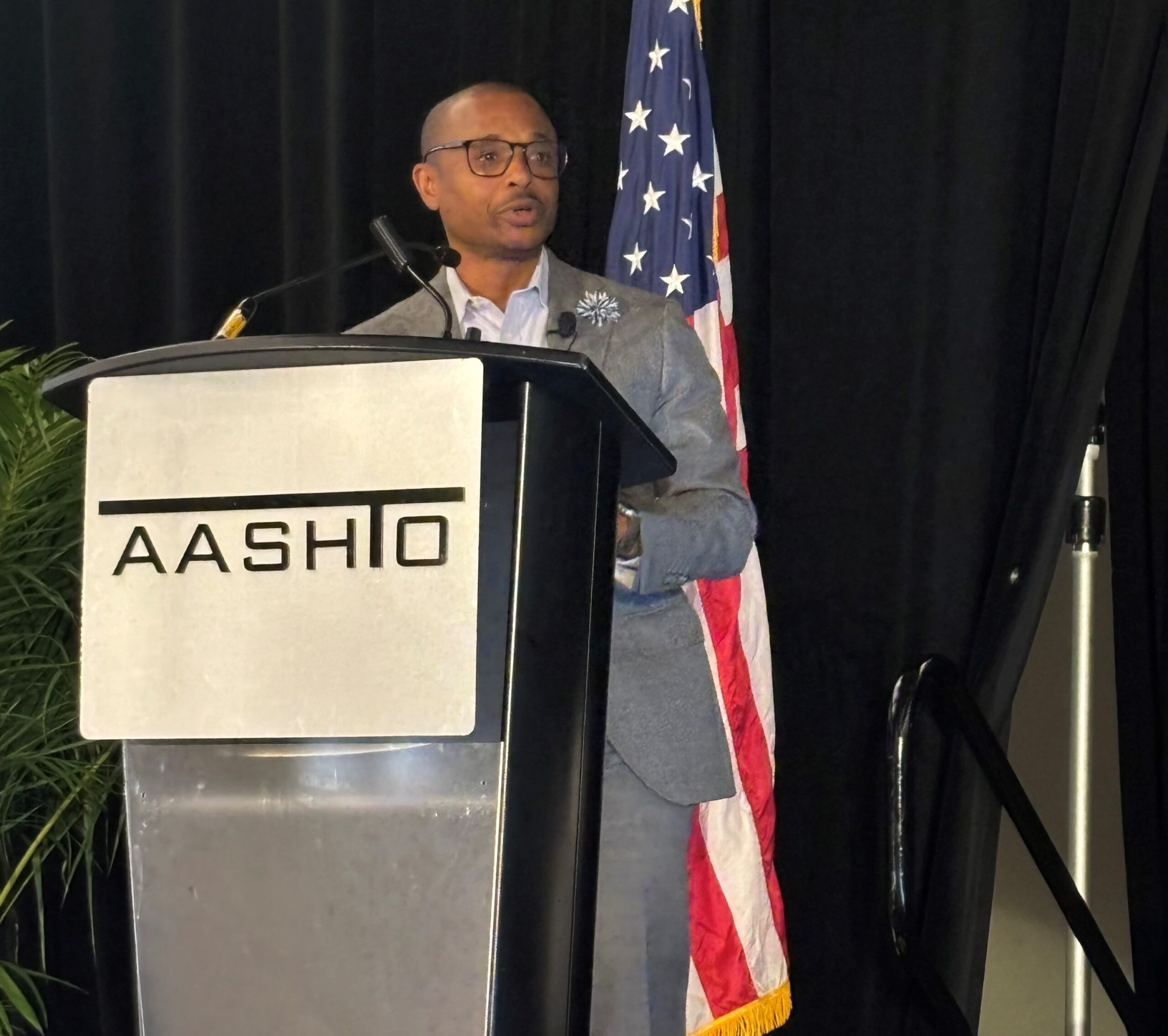 Man in a gray suit speaking at a podium with "AASHTO" logo; American flag in the background.