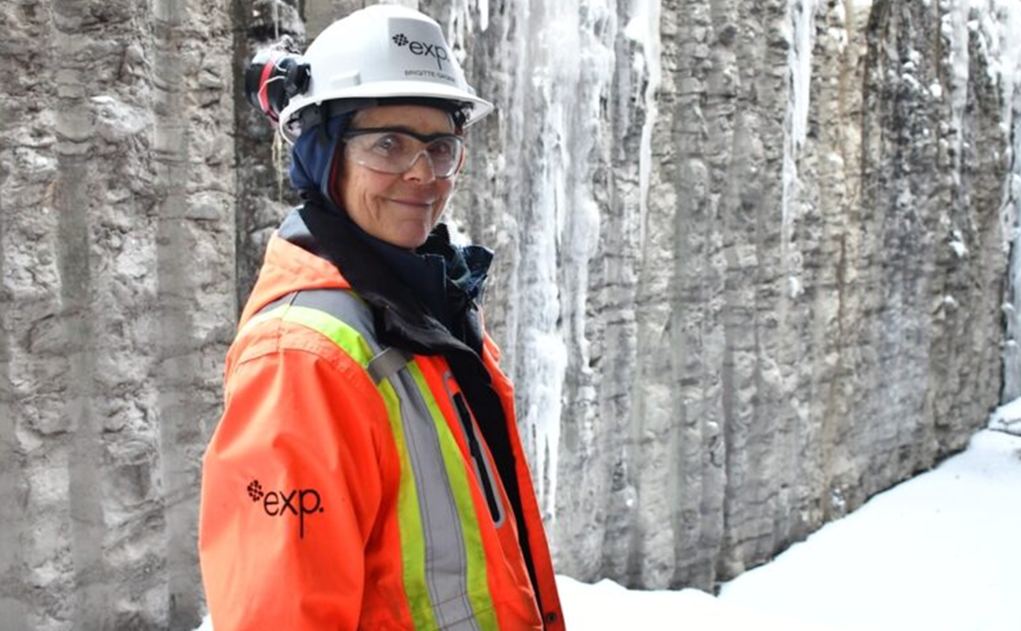 Une personne portant un équipement de sécurité, notamment un casque et une veste orange, se tient dans un environnement enneigé et glacé avec un mur de pierre en arrière-plan.