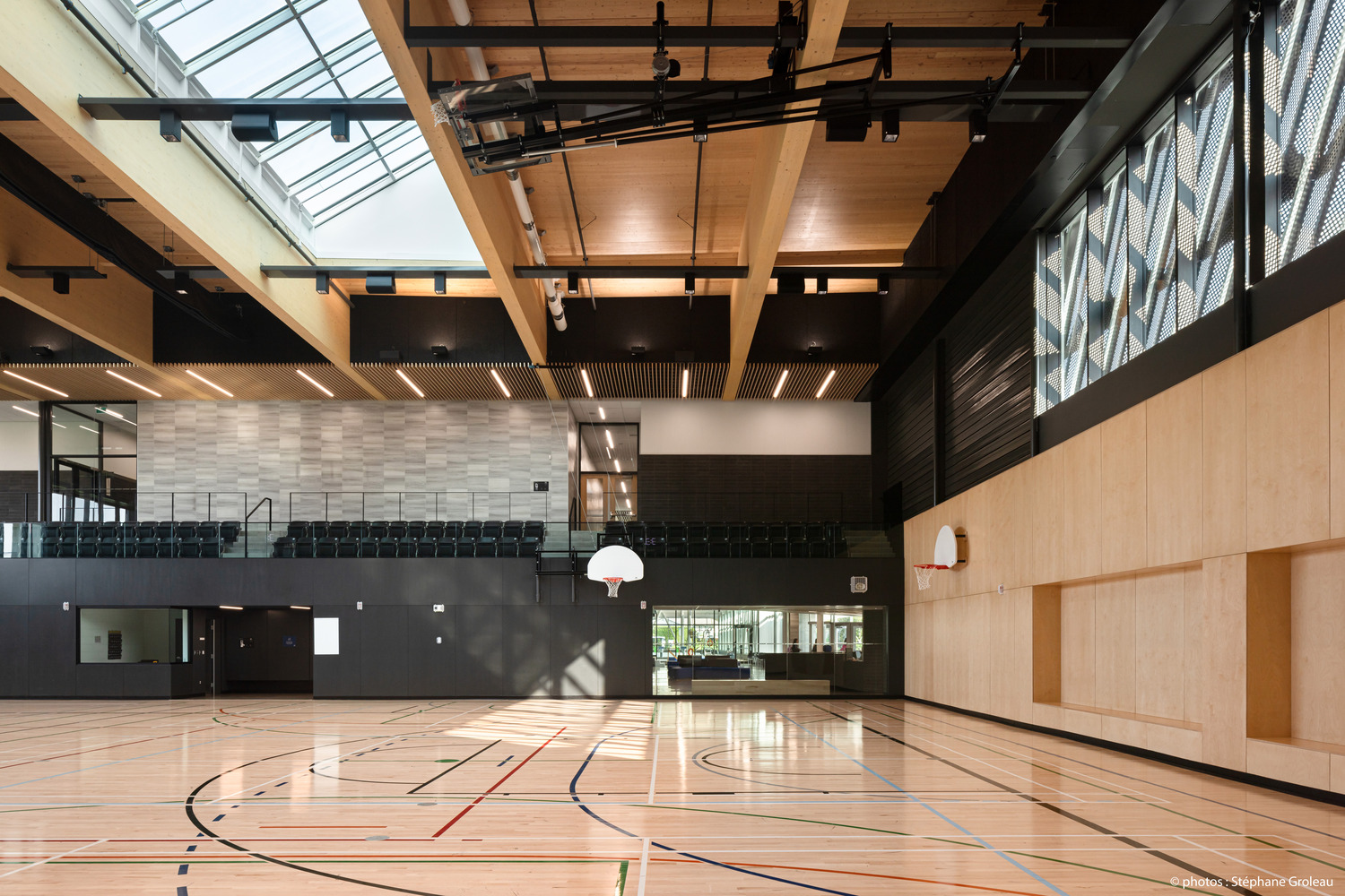 Intérieur d'un gymnase moderne avec un plancher en bois, deux paniers de basket et un coin salon. La lumière naturelle pénètre à travers de grandes fenêtres et des puits de lumière.