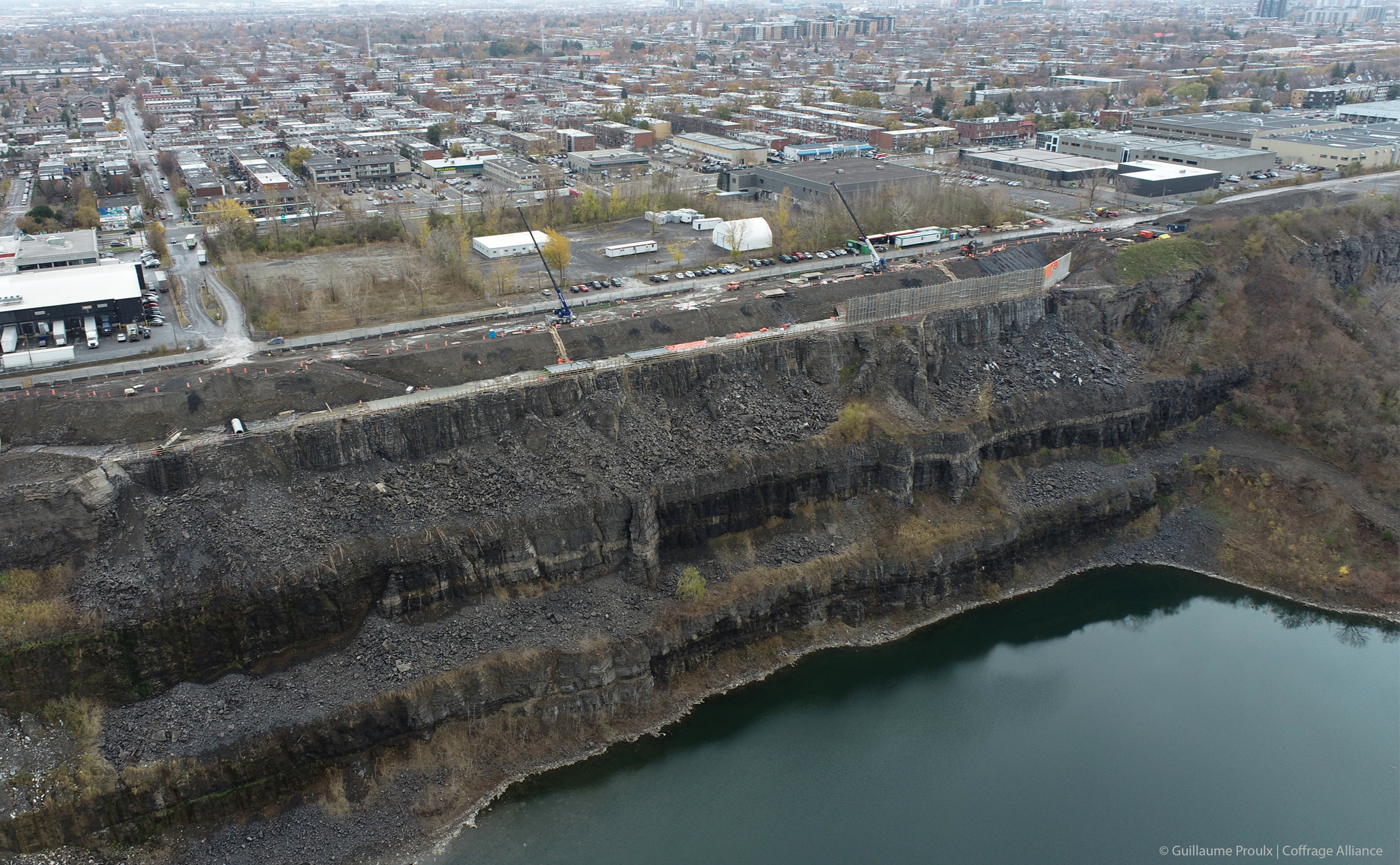 Vue aérienne d'une grande carrière à côté d'un plan d'eau, entourée de bâtiments industriels et d'un paysage urbain en arrière-plan.