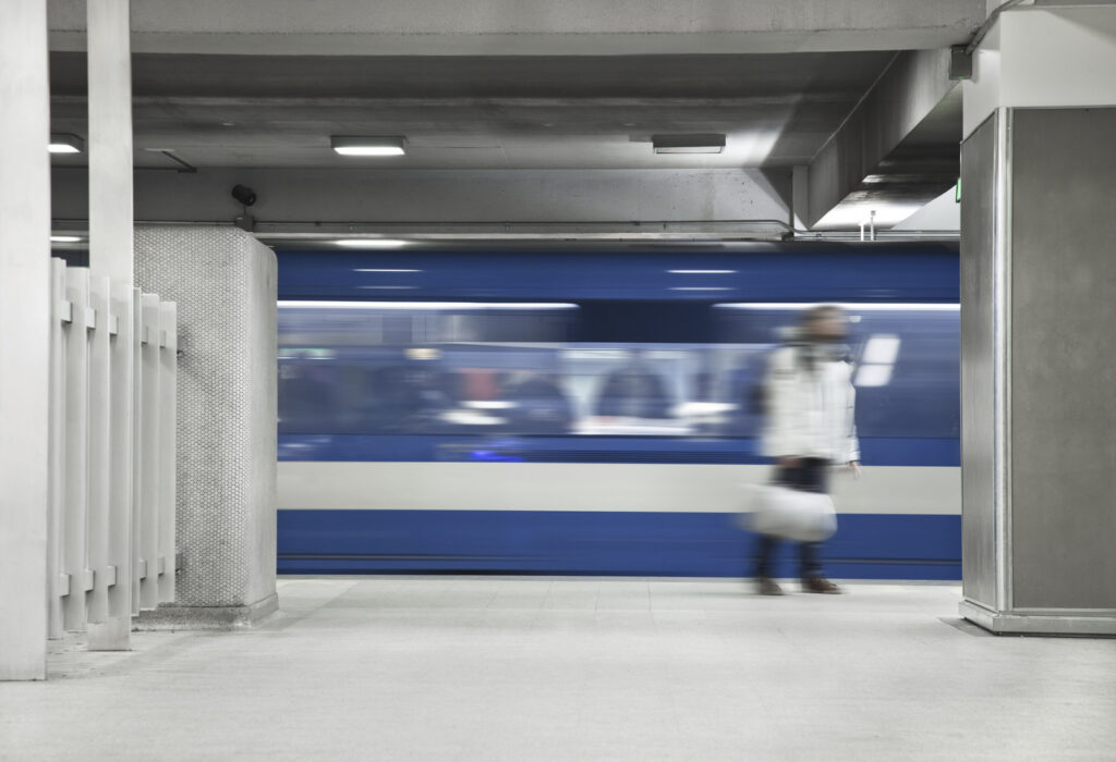 A blue train moves swiftly through an underground station as a person, slightly blurred, stands nearby carrying a bag.