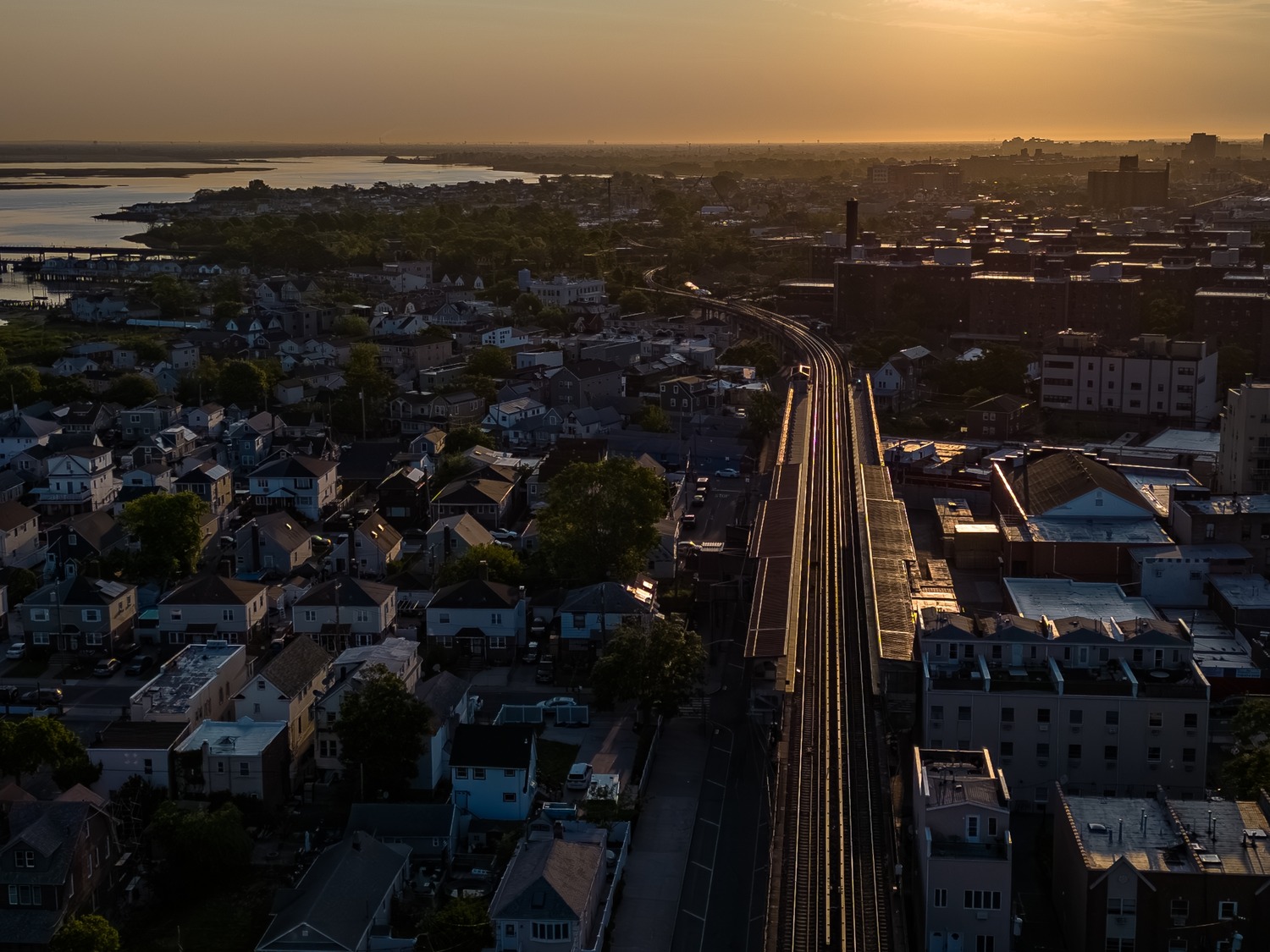 Vue aérienne d'un quartier résidentiel traversé par une voie ferrée menant vers l'eau au coucher du soleil.