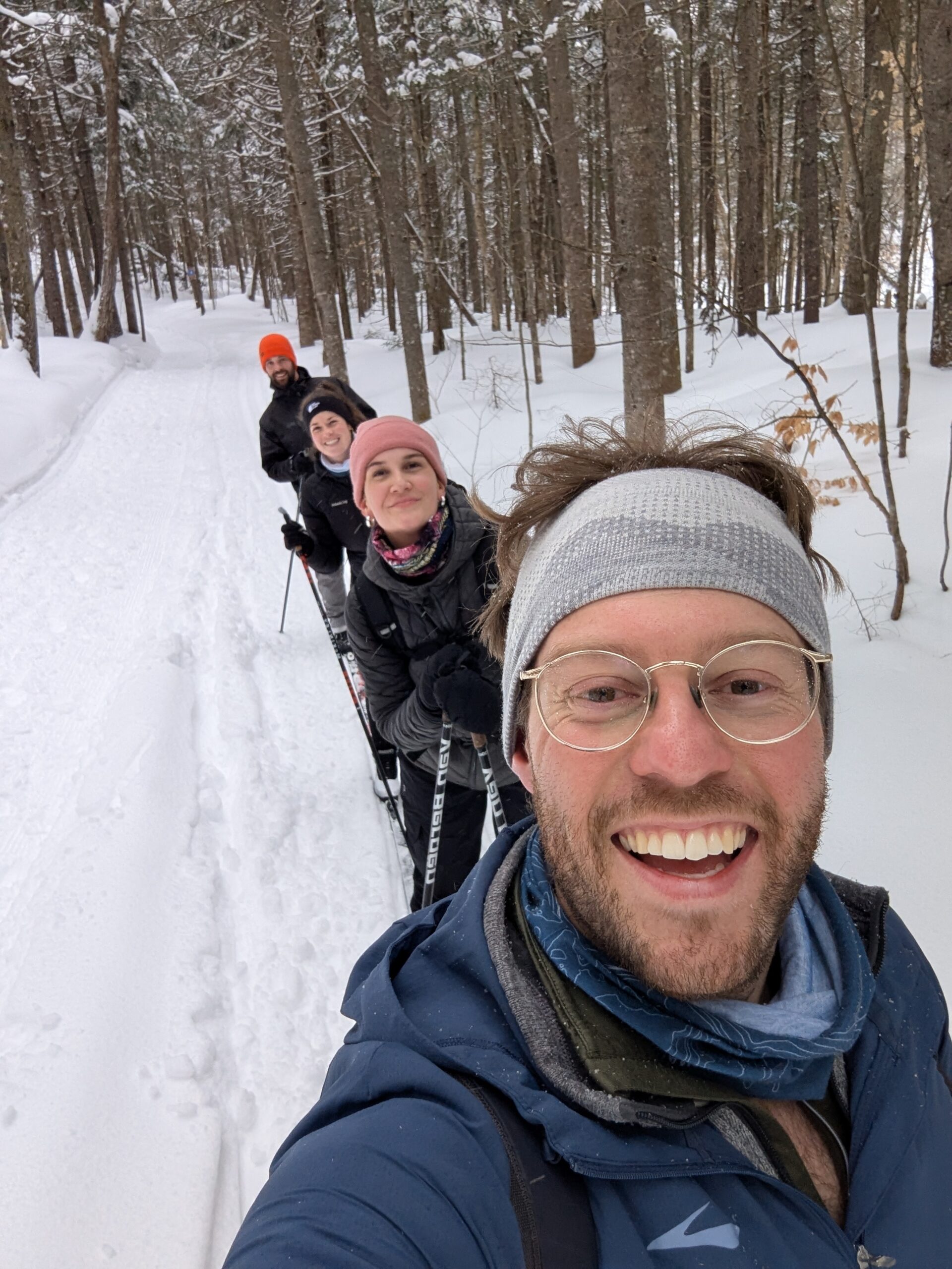 Quatre personnes en vêtements d'hiver sourient en skiant sur un sentier enneigé dans une forêt.
