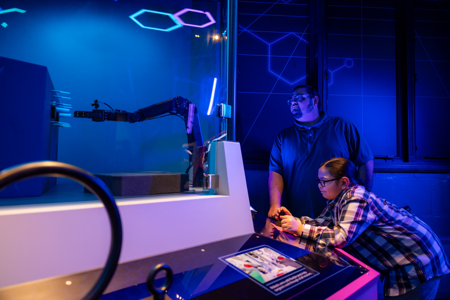 A man and a young girl operate a robotic arm behind glass in a dimly lit, blue-illuminated science exhibit.