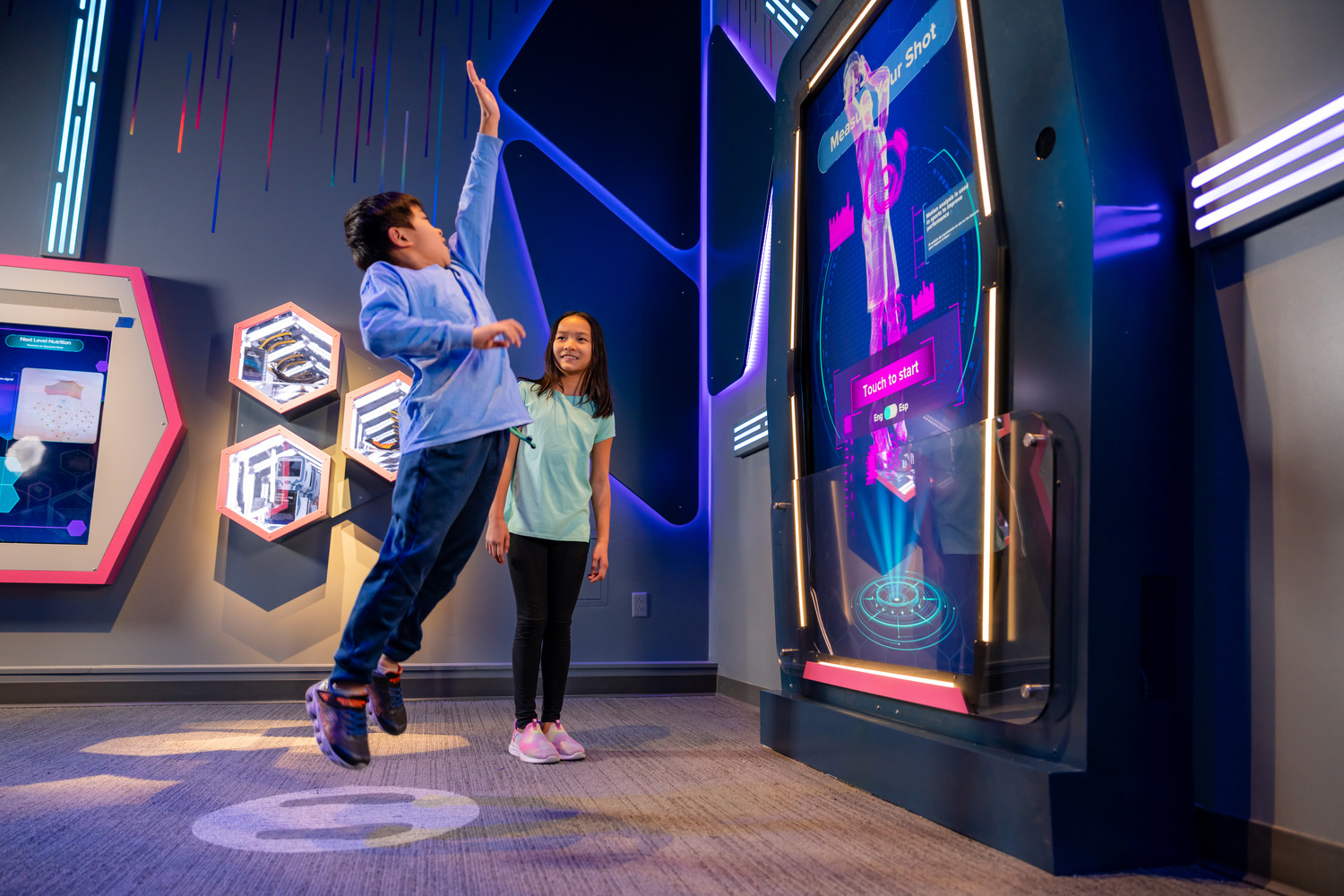 A boy jumps to touch a large interactive screen while a girl watches, both inside a modern, tech-themed exhibit space.