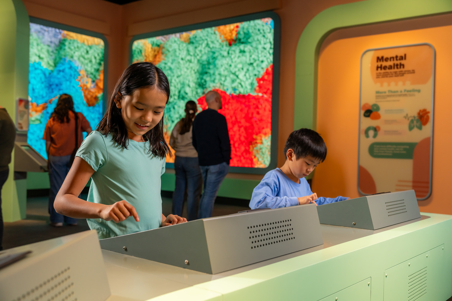 Two children interact with hands-on exhibits in a science museum, while adults observe displays and colorful walls in the background.