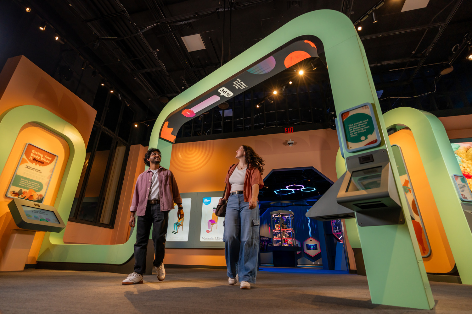 Two people walk through a brightly lit, interactive exhibit with colorful arches and digital displays in a modern museum setting.