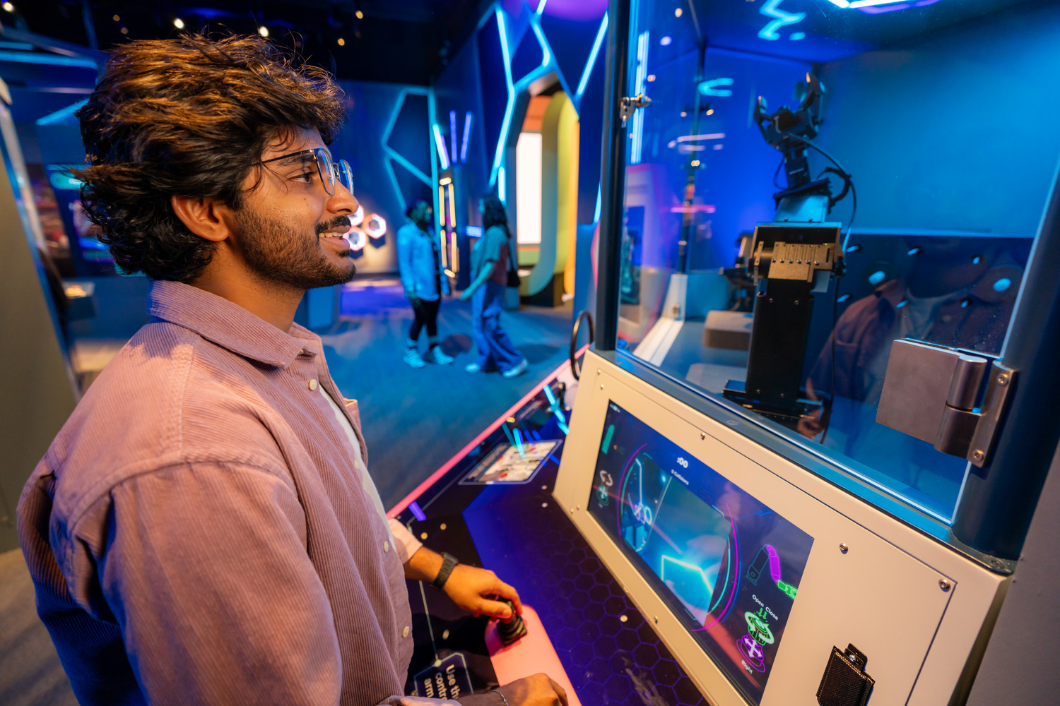 A man in a purple shirt operates a robotic arm at an interactive exhibit with neon lighting and digital displays.