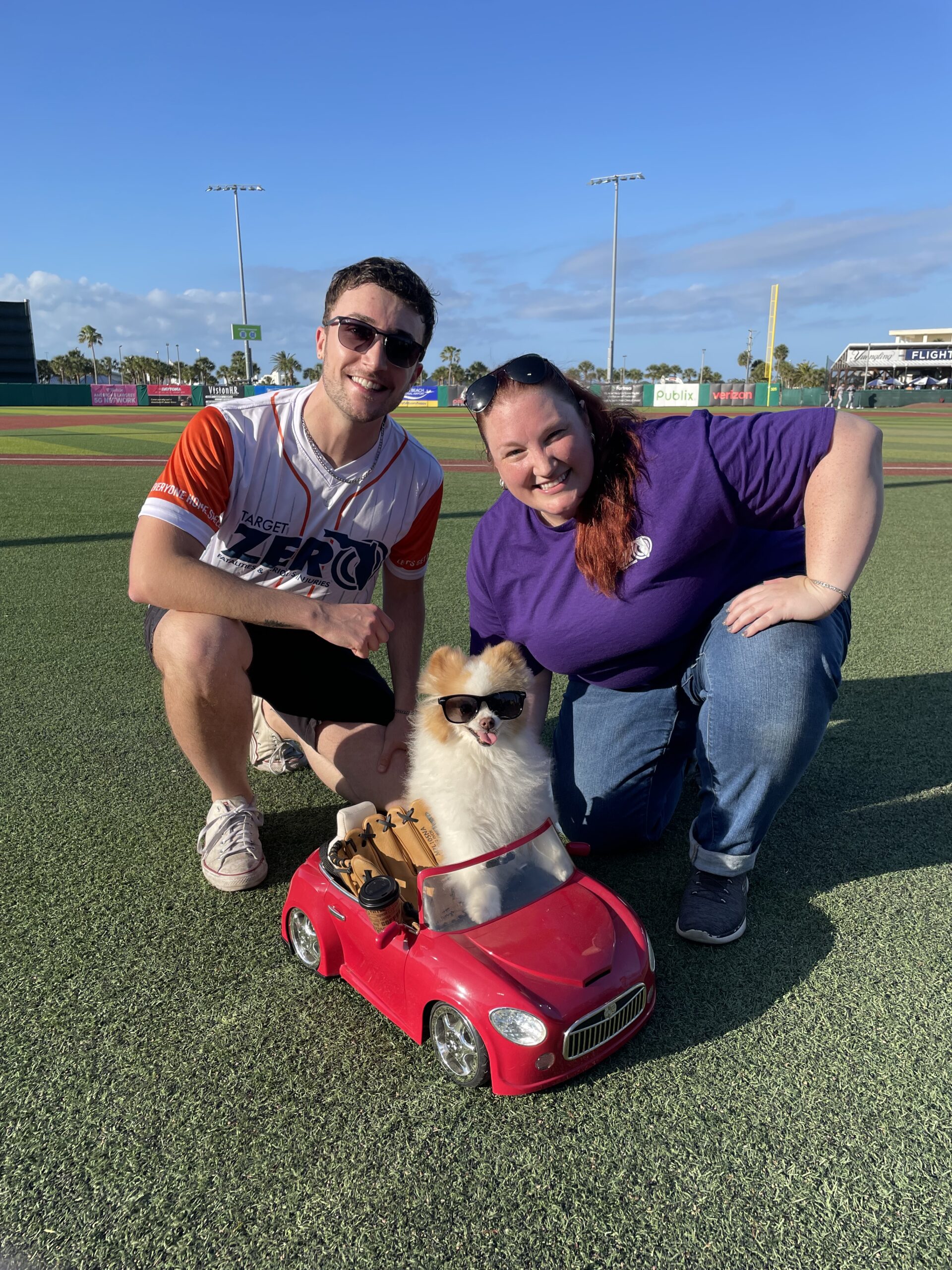 Two people pose on a baseball field with a small dog wearing sunglasses, sitting in a red toy convertible car.
