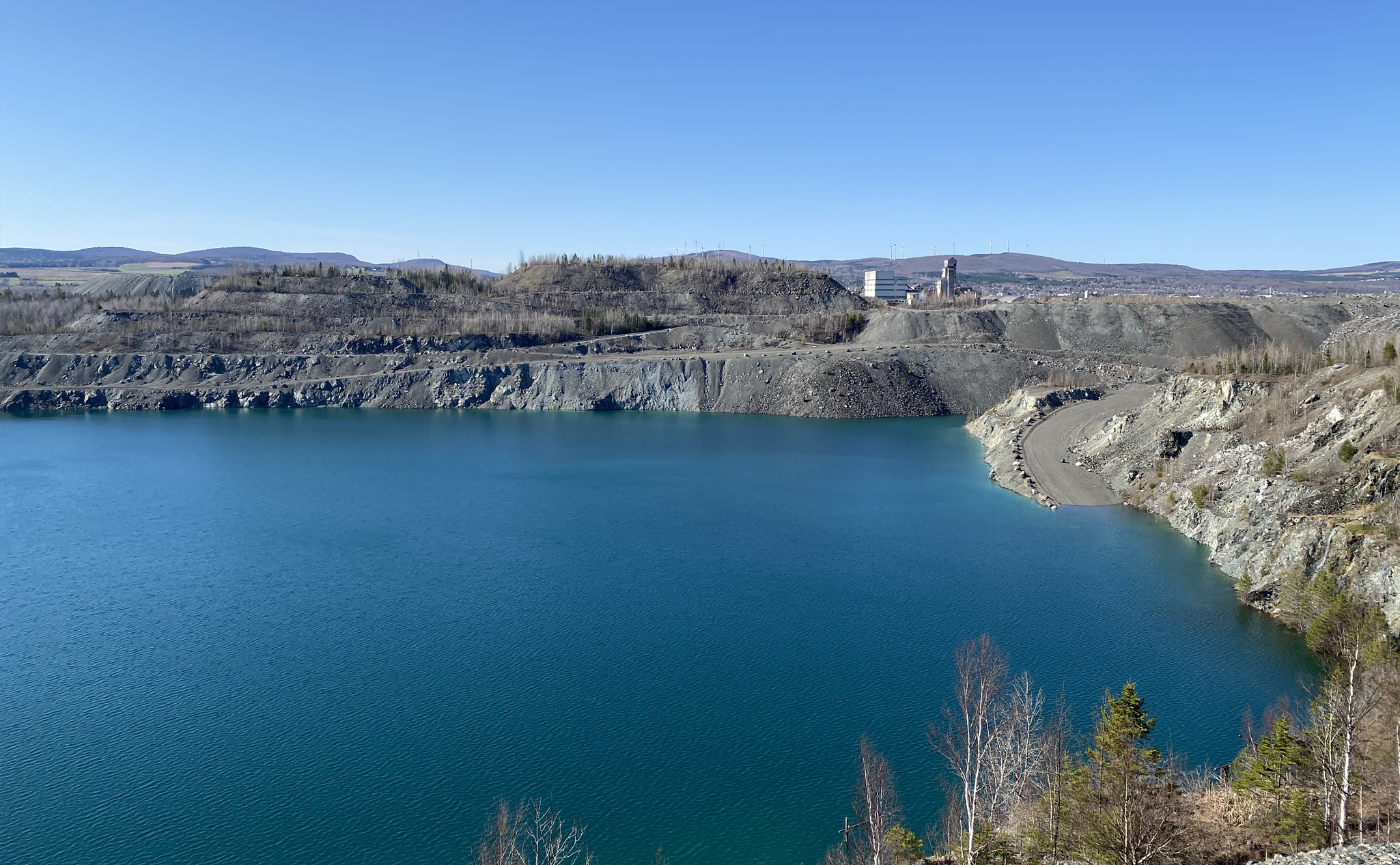 Un grand lac bleu occupe le premier plan, bordé de terrains rocheux et d'arbres clairsemés, avec une installation industrielle visible au loin sous un ciel clair.