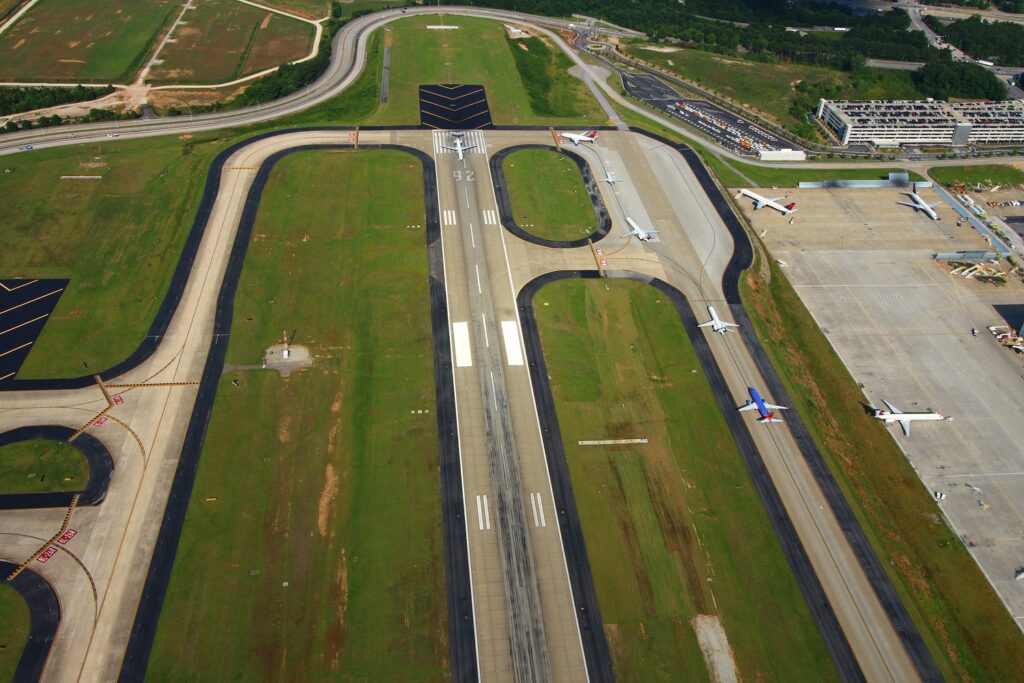 Aerial view of an airport runway with several planes taxiing and parked near the terminal, surrounded by green grass and roads—a scene reminiscent of travel hubs featured at the AASHTO Annual Meeting.