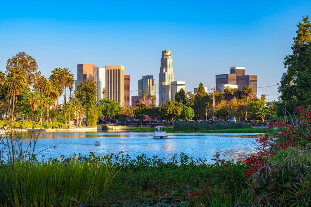Downtown Los Angeles skyline viewed from a park with a lake in the foreground, lush greenery, palm trees, and a boat on the water under a clear blue sky—capturing the vibrant spirit of Mobility 21 in an urban oasis.