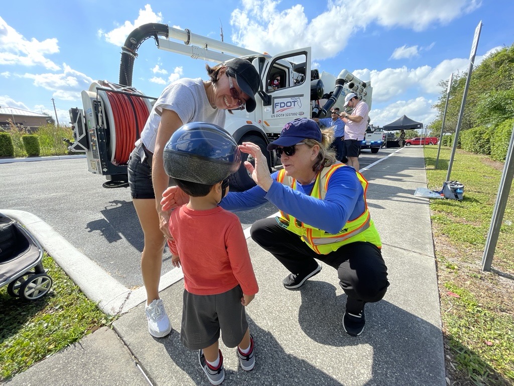 A woman in a safety vest adjusts a helmet on a young boy while another woman watches, with an FDOT truck and equipment in the background on a sunny day.
