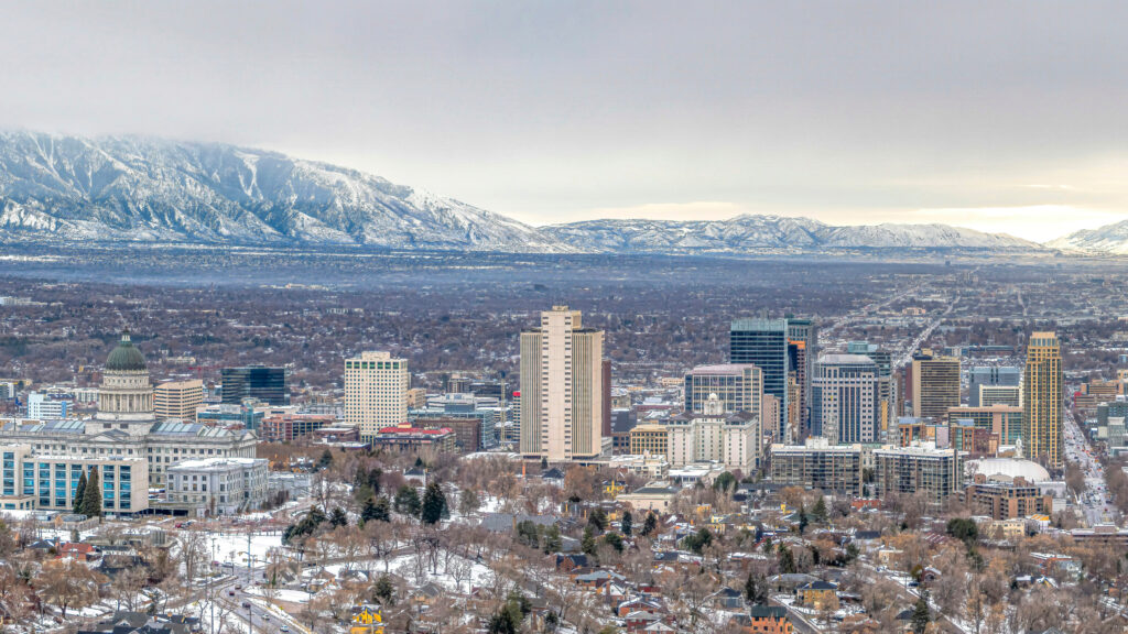 A cityscape with tall buildings, snow-covered rooftops, and distant mountains partially shrouded in clouds under a gray sky, reflecting a commitment to sustainability through innovative urban design and commissioning practices.