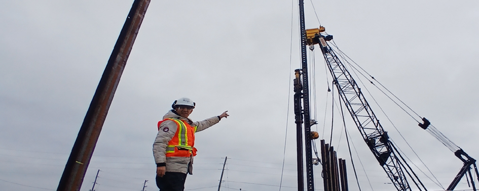 A construction worker in safety gear points upward toward a tall pile-driving crane at an outdoor construction site under a cloudy sky.
