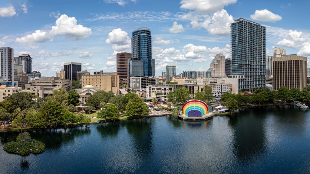 Downtown city skyline with modern buildings known for their sustainability, a lake in the foreground, and a colorful rainbow-shaped amphitheater on the shoreline under a partly cloudy sky.