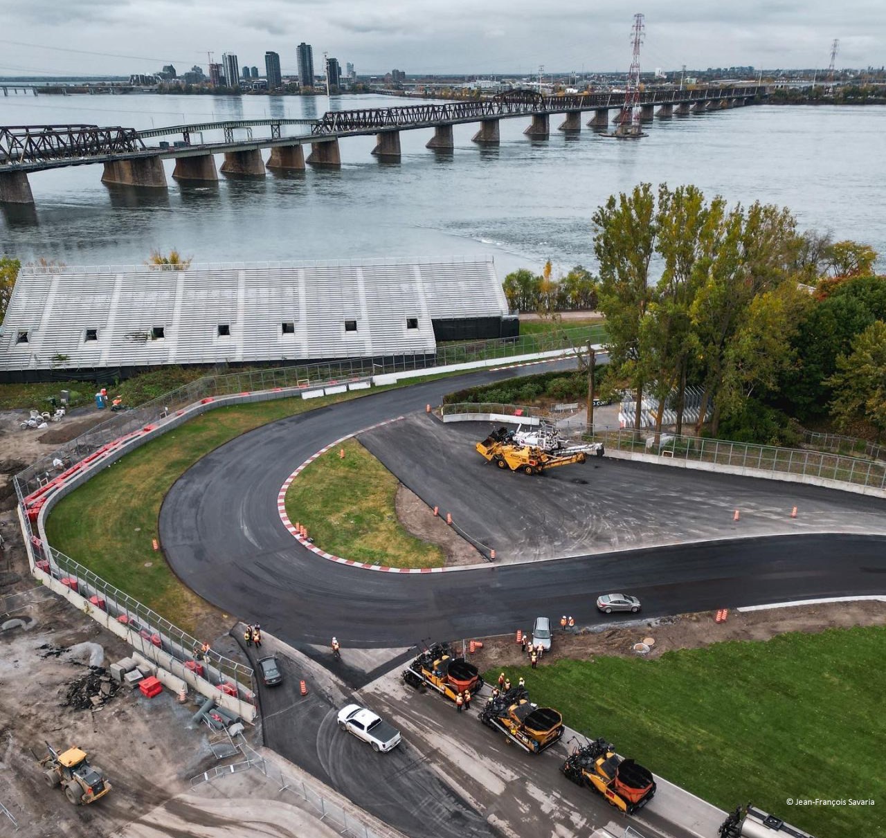 Vue aérienne d'une construction routière sur une piste courbe près d'une rivière, avec plusieurs véhicules de construction et un pont en arrière-plan.