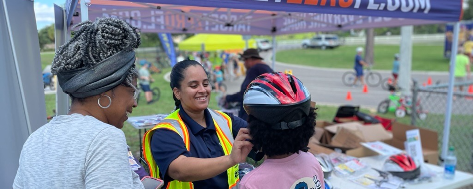 A safety worker fits a child with a bike helmet at an outdoor event, while another adult stands nearby under a canopy.