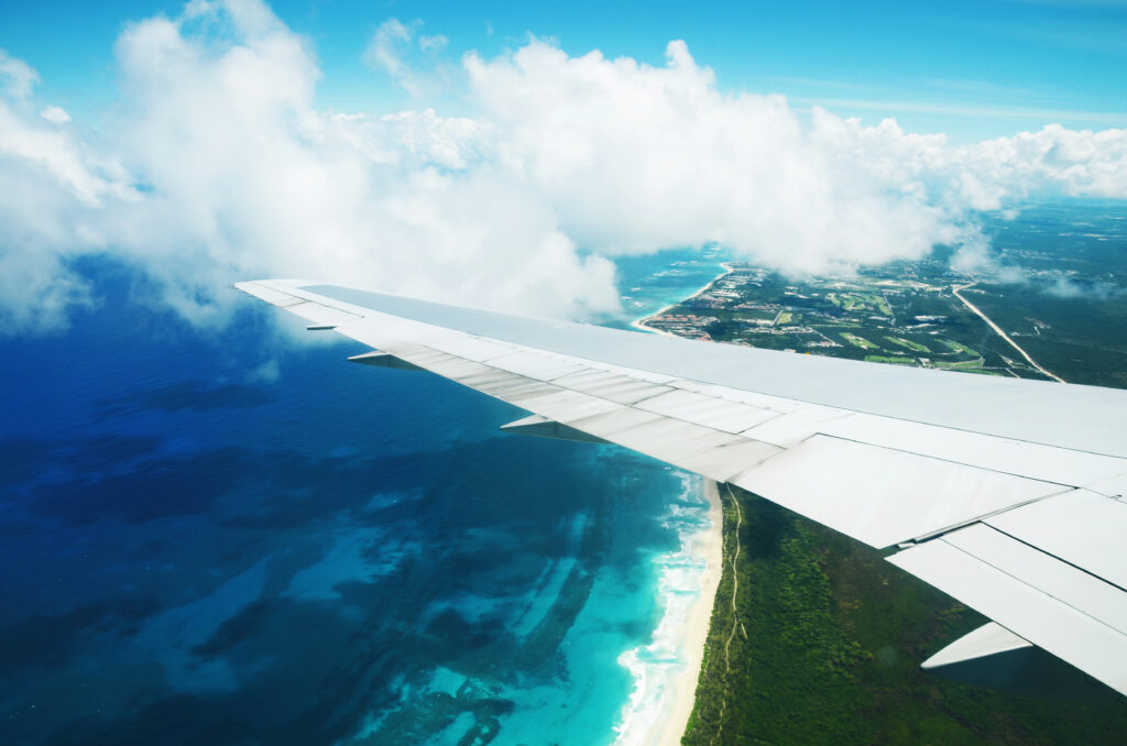 View from an airplane window showing a wing over turquoise ocean water, white sandy beach, and coastal landscape with some clouds in the sky en route to the LAMAT Airport Summit III.