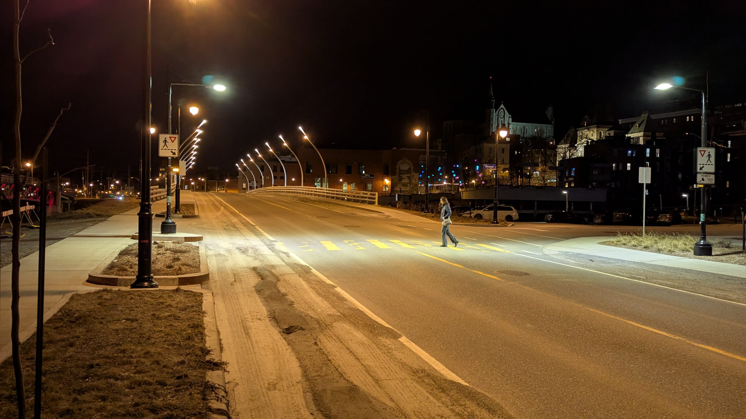 Une personne traverse une rue vide et bien éclairée la nuit près d'un pont, avec des bâtiments et des lampadaires visibles en arrière-plan.