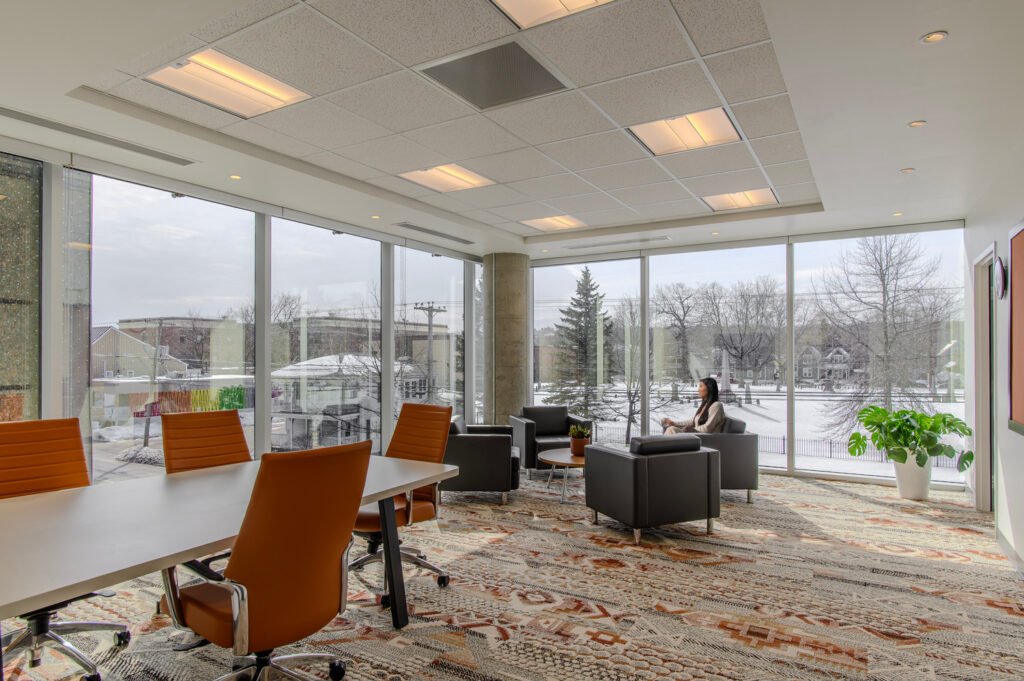 Modern office space with floor-to-ceiling windows, orange chairs around a conference table, seating area with armchairs, and a person sitting by the window overlooking a snowy landscape.
