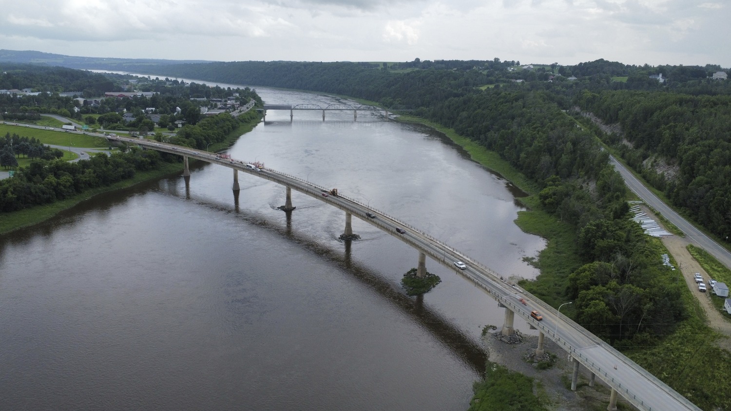 Aerial view of a long bridge crossing a wide river with cars and trucks, surrounded by green trees and distant hills under a cloudy sky.
