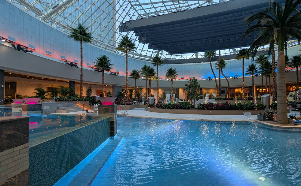 Indoor pool with clear blue water, artificial palm trees, and a glass dome ceiling. Ambient lighting and plants line the poolside area.