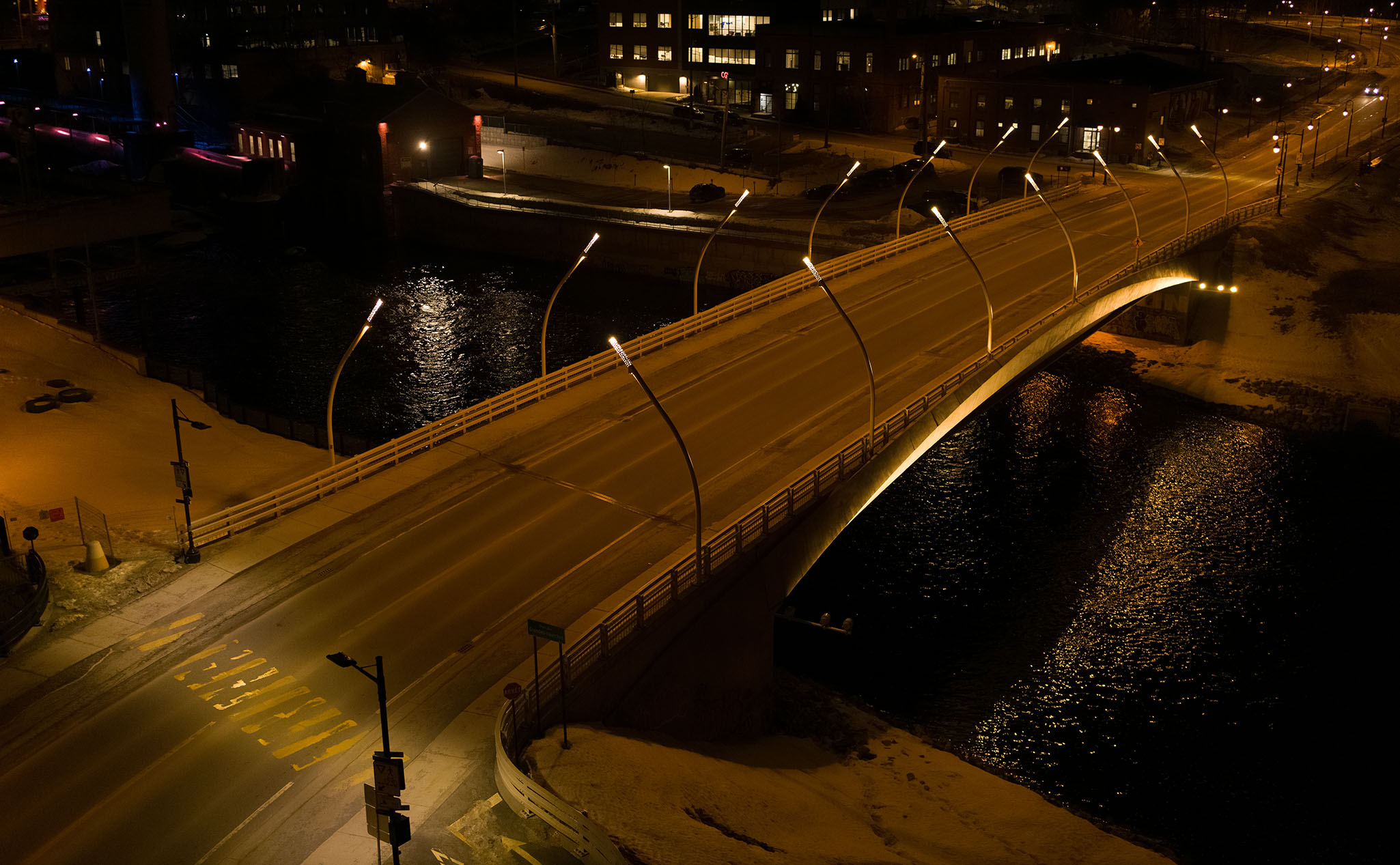 Un pont urbain éclairé par des lampadaires la nuit traverse une rivière, avec de la neige et des bâtiments visibles dans les environs.