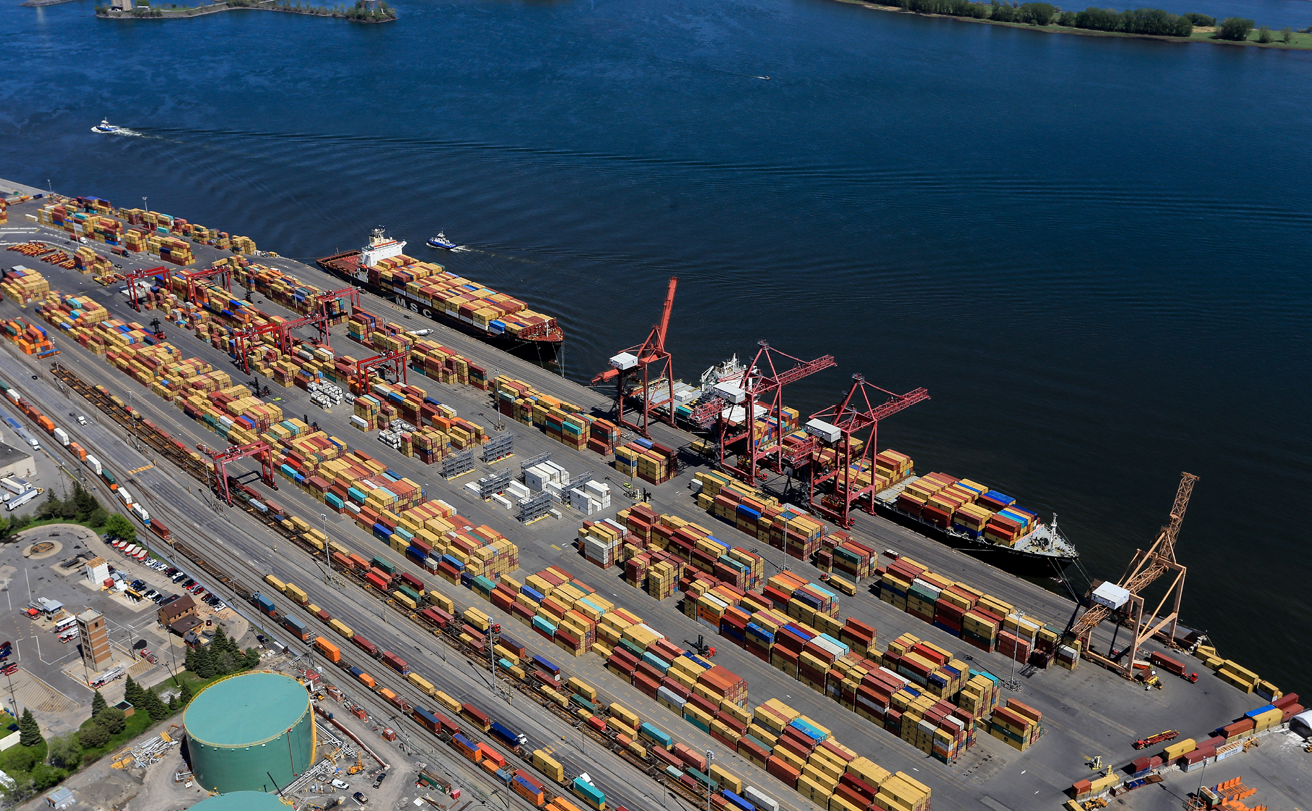 Vue aérienne d'un port d'expédition avec des conteneurs d'expédition empilés, des grues de chargement et deux grands porte-conteneurs amarrés au bord de l'eau.