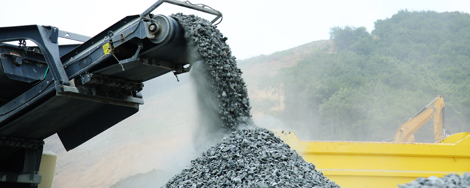 A TARBA conveyor belt deposits crushed stone onto a large pile, with a yellow industrial vehicle and hills visible in the background.