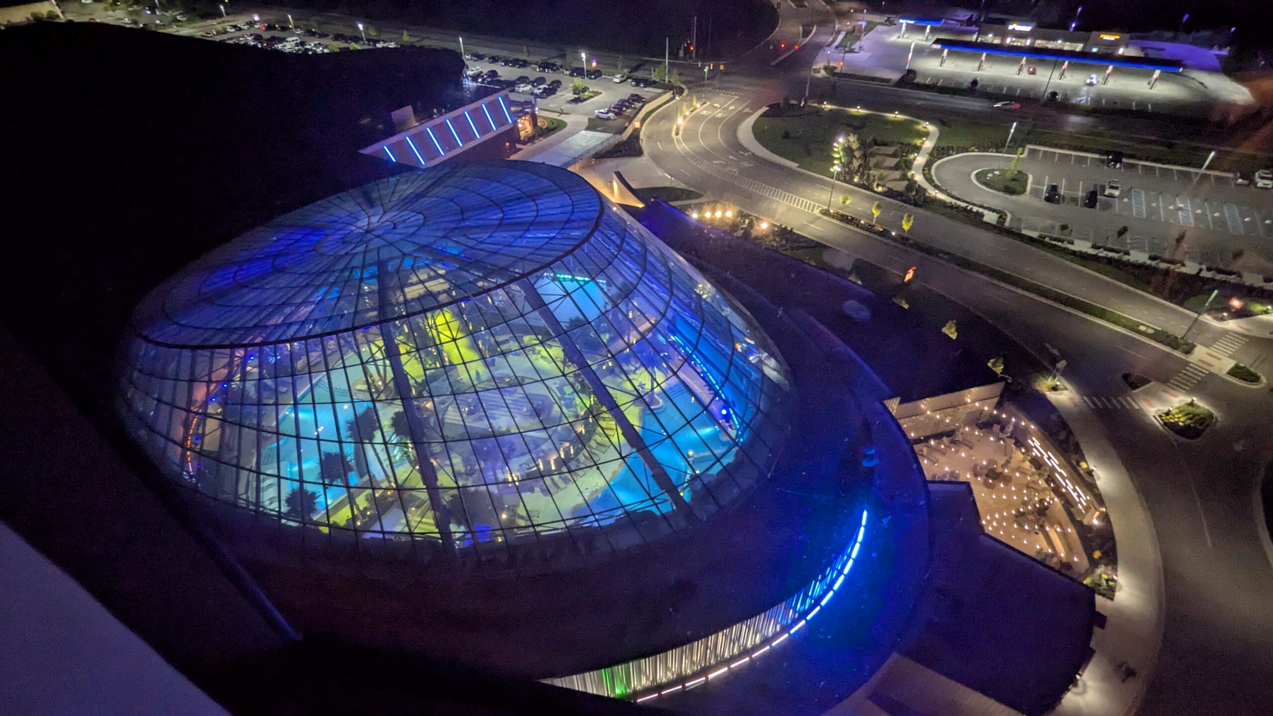 Aerial night view of a large glass-domed building with blue and yellow lighting, next to roads, parking lots, and other illuminated structures.