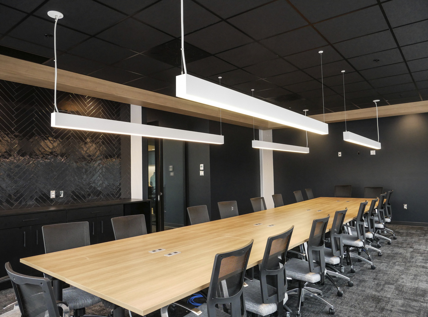 Modern conference room with a long wooden table, several office chairs, overhead rectangular lights, and a black accent wall.