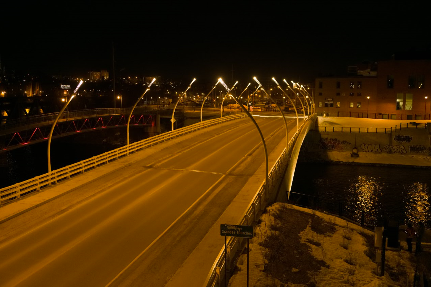 Un pont bien éclairé sur une rivière la nuit avec des routes vides, des bâtiments à proximité et des graffitis sur un mur au bord de l'eau.