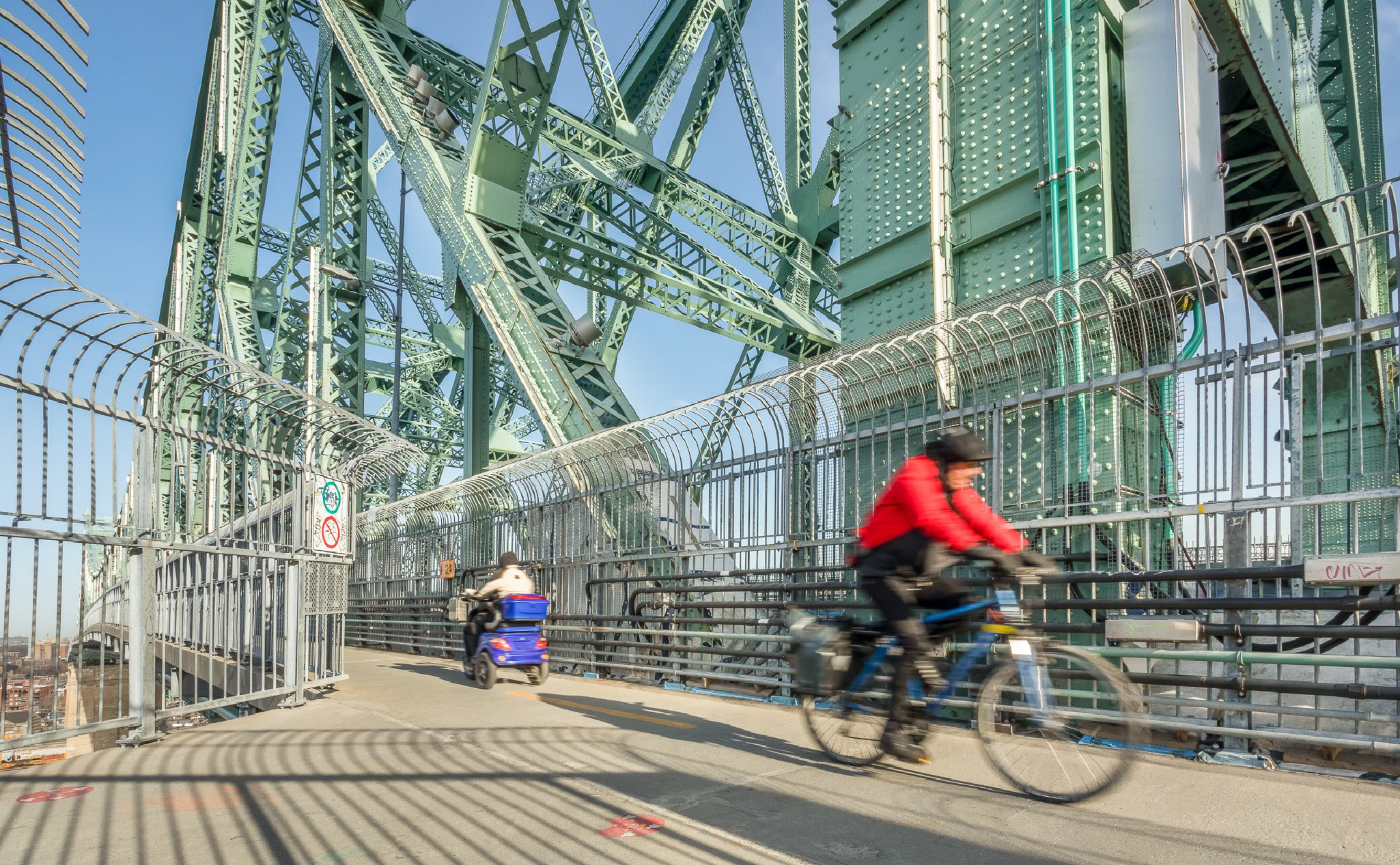 Une personne à vélo et une autre sur un scooter de mobilité traversent un pont clôturé avec de grands supports en acier vert en arrière-plan.