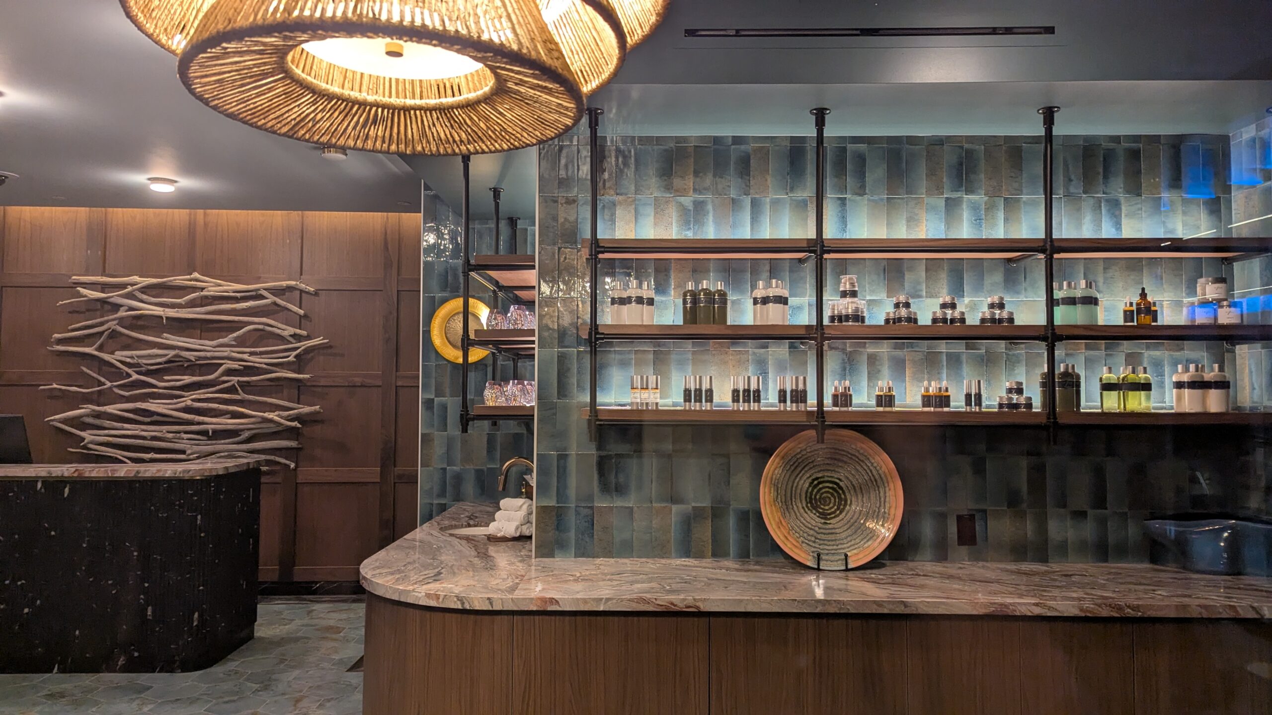 Modern spa reception area with a marble counter, decorative plates, shelves holding bottles, and a wood art piece on the back wall.
