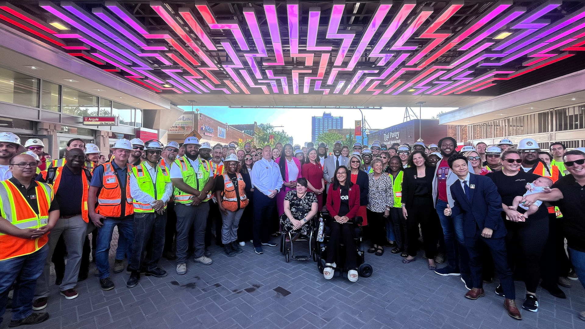 A large group of people, including construction workers in safety vests and others in business attire, pose together under a ceiling with colorful, geometric lighting for the Red and Purple Modernization Phase One Project.