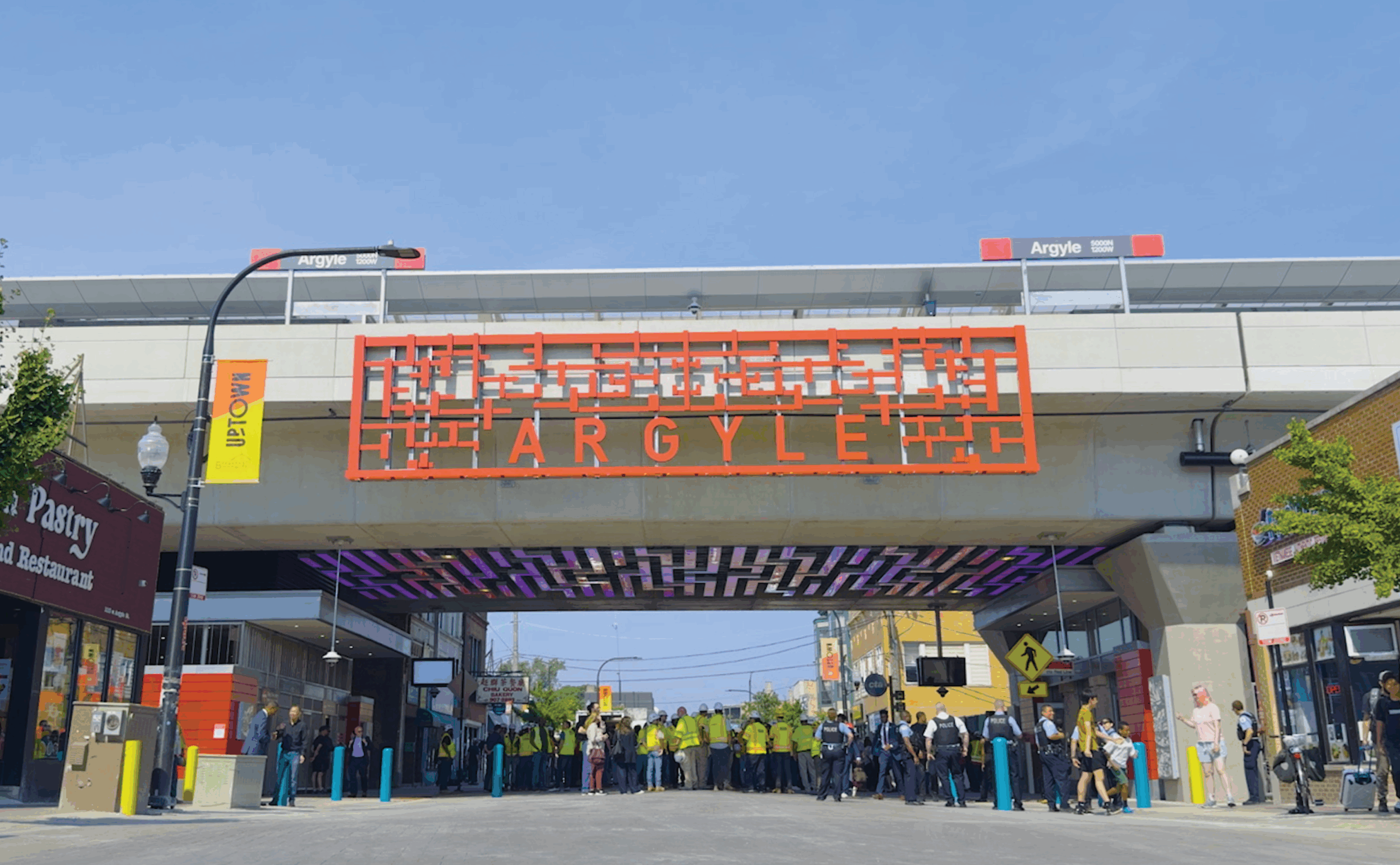 Un groupe de personnes se rassemble sous le viaduc de la station CTA Argyle, qui comporte un panneau géométrique rouge et les devantures des magasins voisins, lors d'une journée ensoleillée.