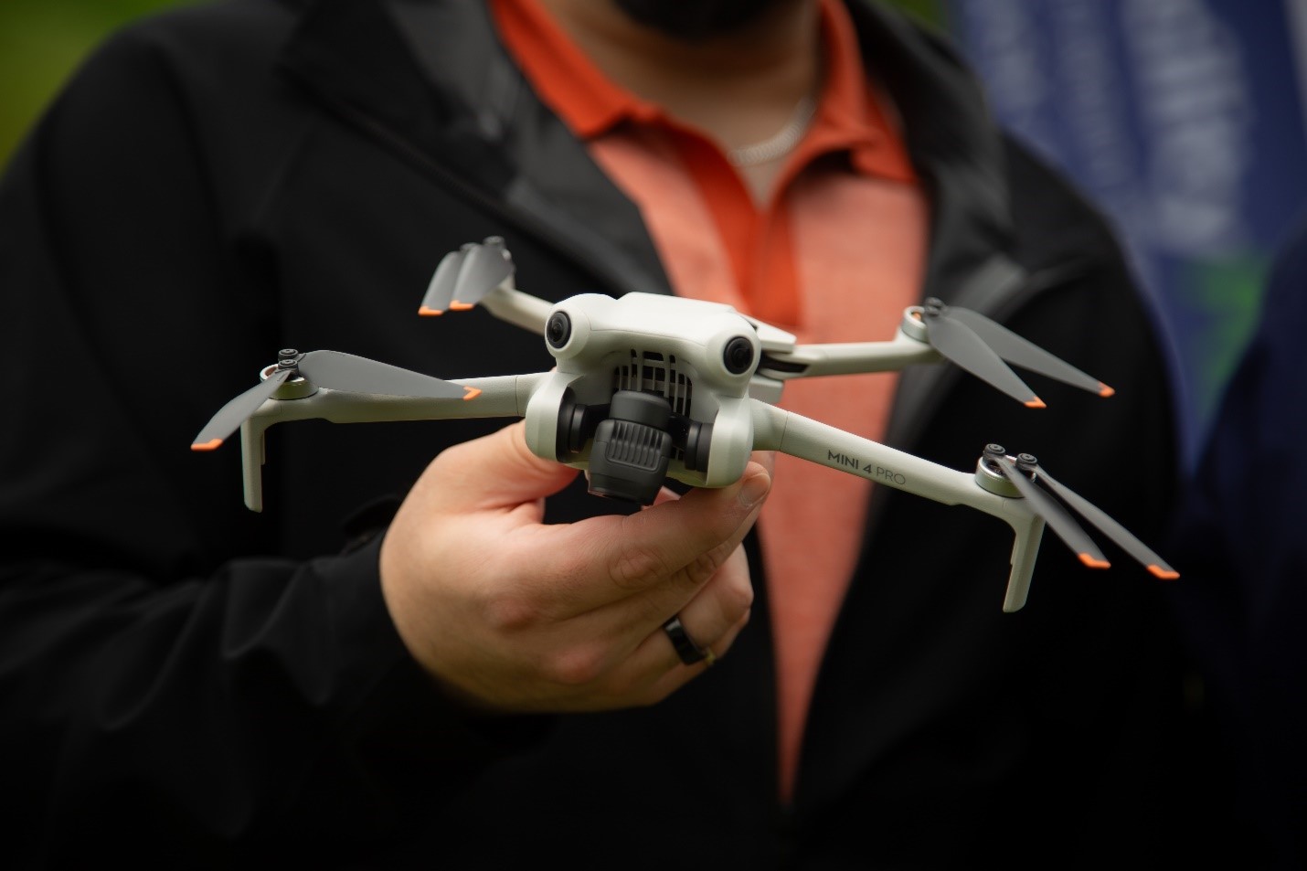 A person holding a small quadcopter drone with a camera, dressed in a black jacket and orange shirt, outdoors.