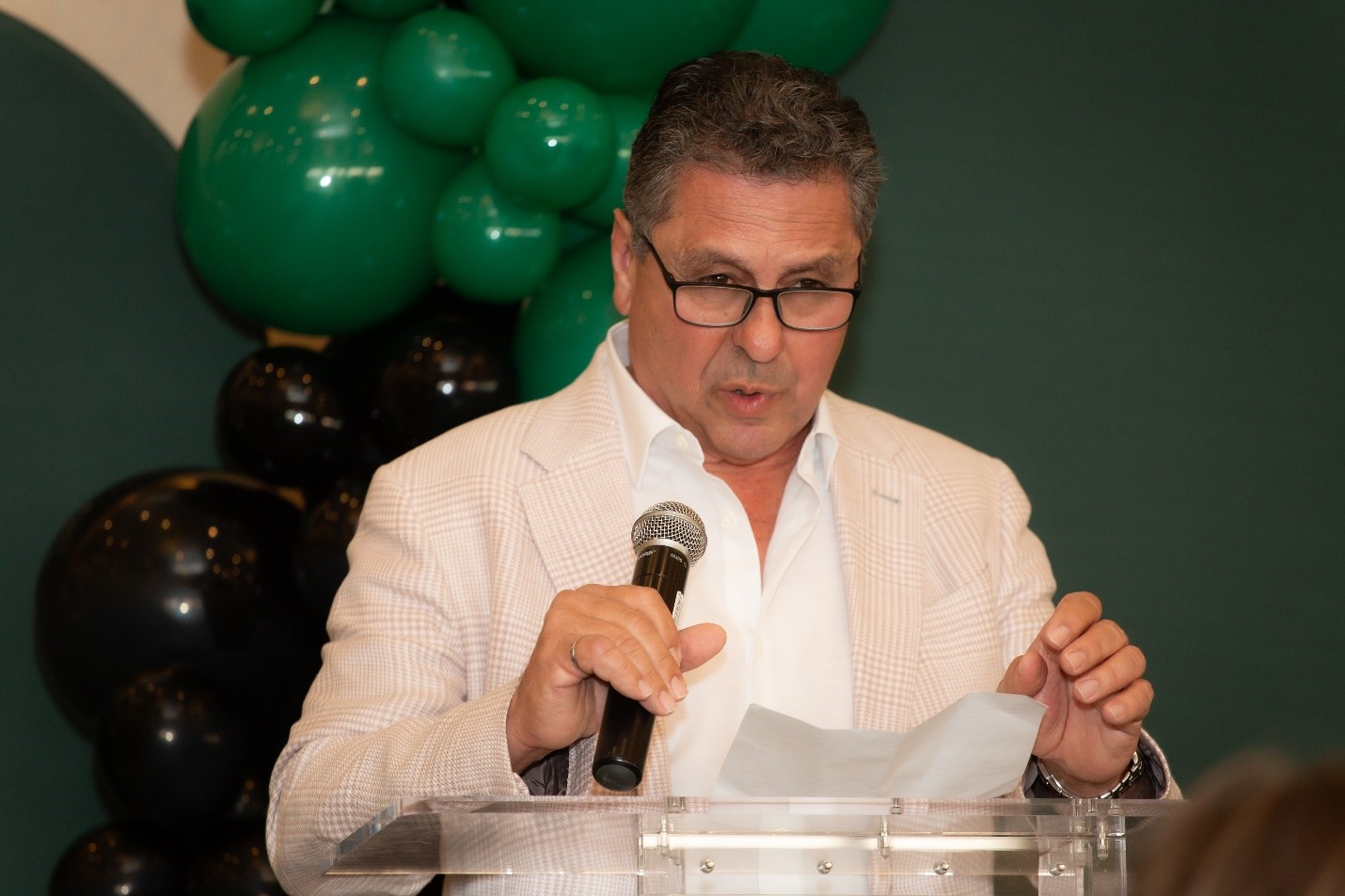 A man in glasses and a light-colored jacket speaks into a microphone while holding a piece of paper at a podium, with green and black balloons in the background.