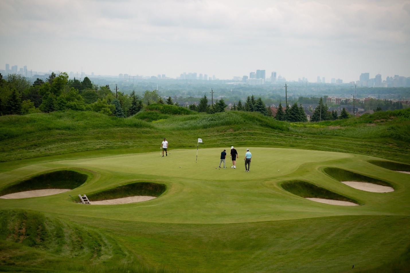 Four people stand on a golf green surrounded by sand bunkers, with a city skyline visible in the background under a cloudy sky.