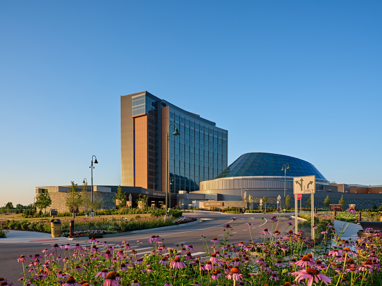 A modern hotel and convention center with a glass dome and tall tower, surrounded by landscaped gardens and purple flowers under a clear blue sky.