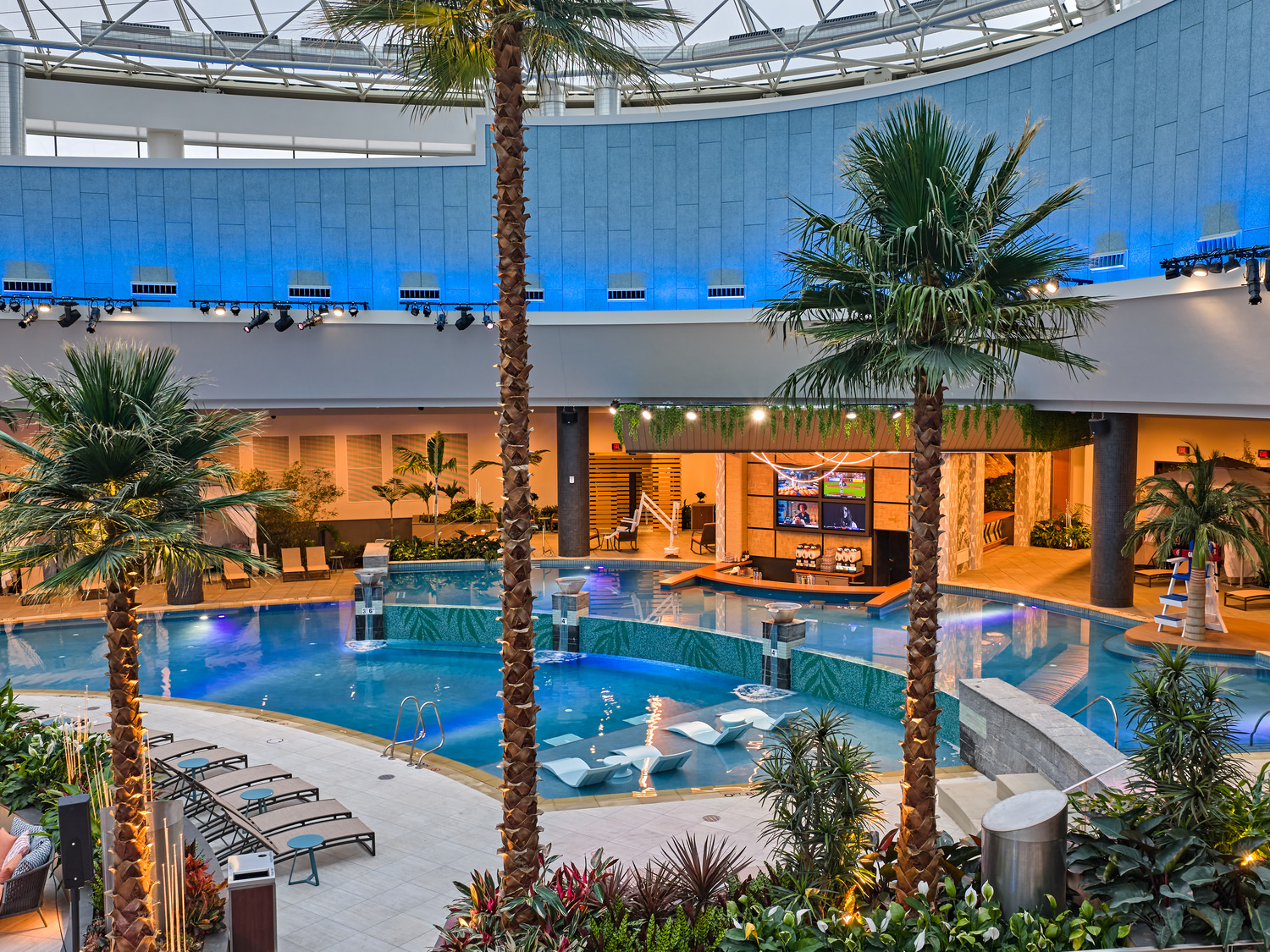 Indoor pool area with palm trees, lounge chairs, a swim-up bar, and tropical plants under a large glass dome ceiling.