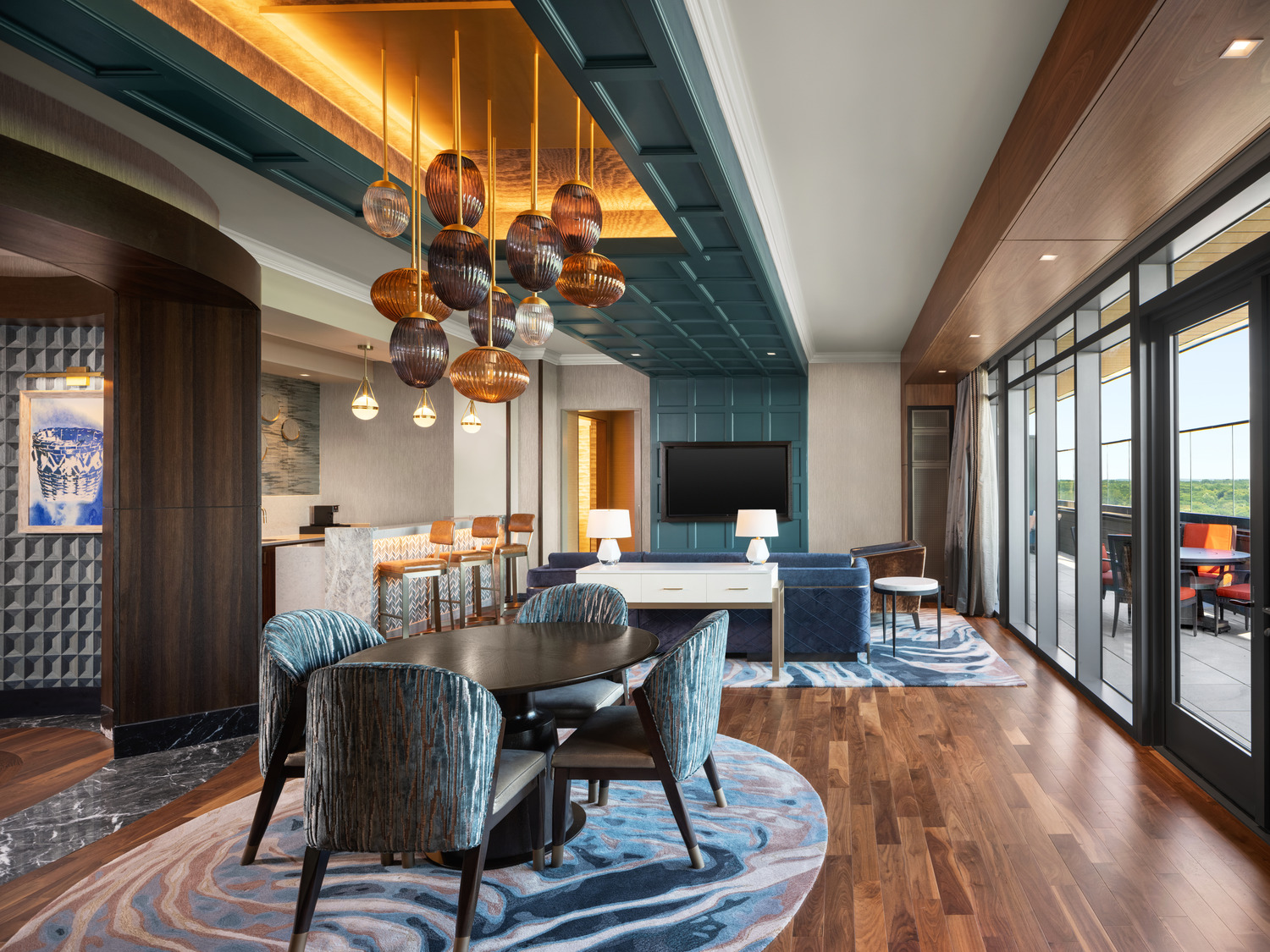 Modern hotel suite with wood floors, blue-patterned rugs and chairs, a central dining table, a lounge area with sofa and TV, large windows, and decorative ceiling lights.
