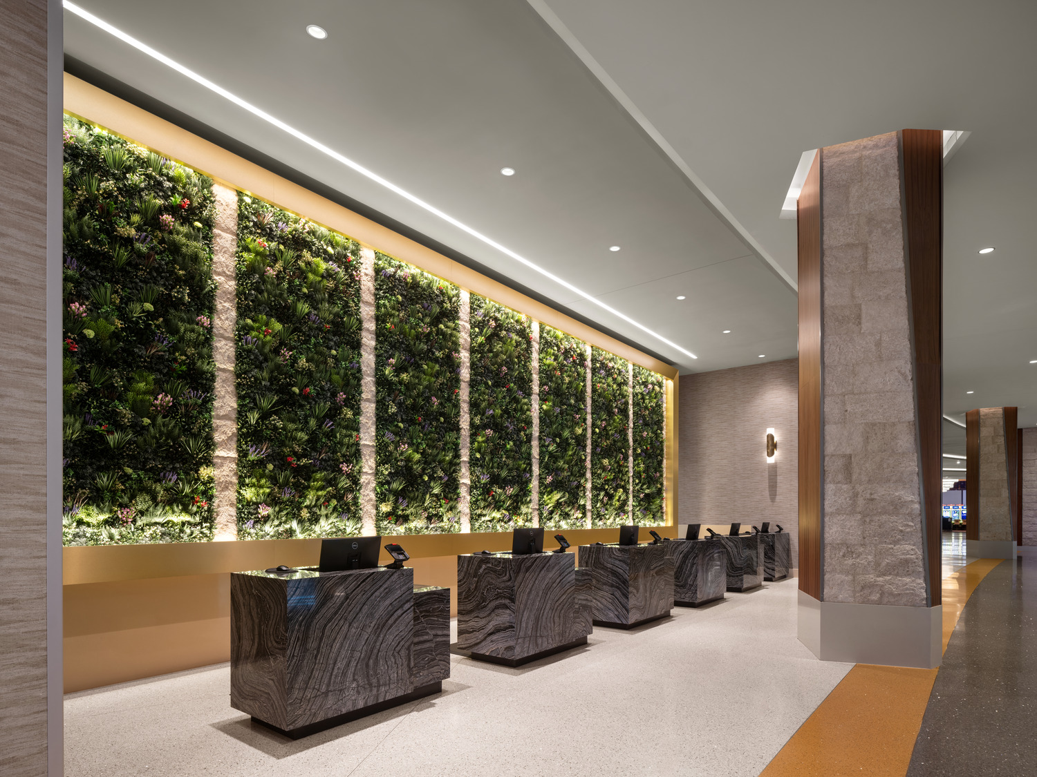 A modern hotel lobby features a green living wall behind several stone reception desks with computers, under bright ceiling lights.