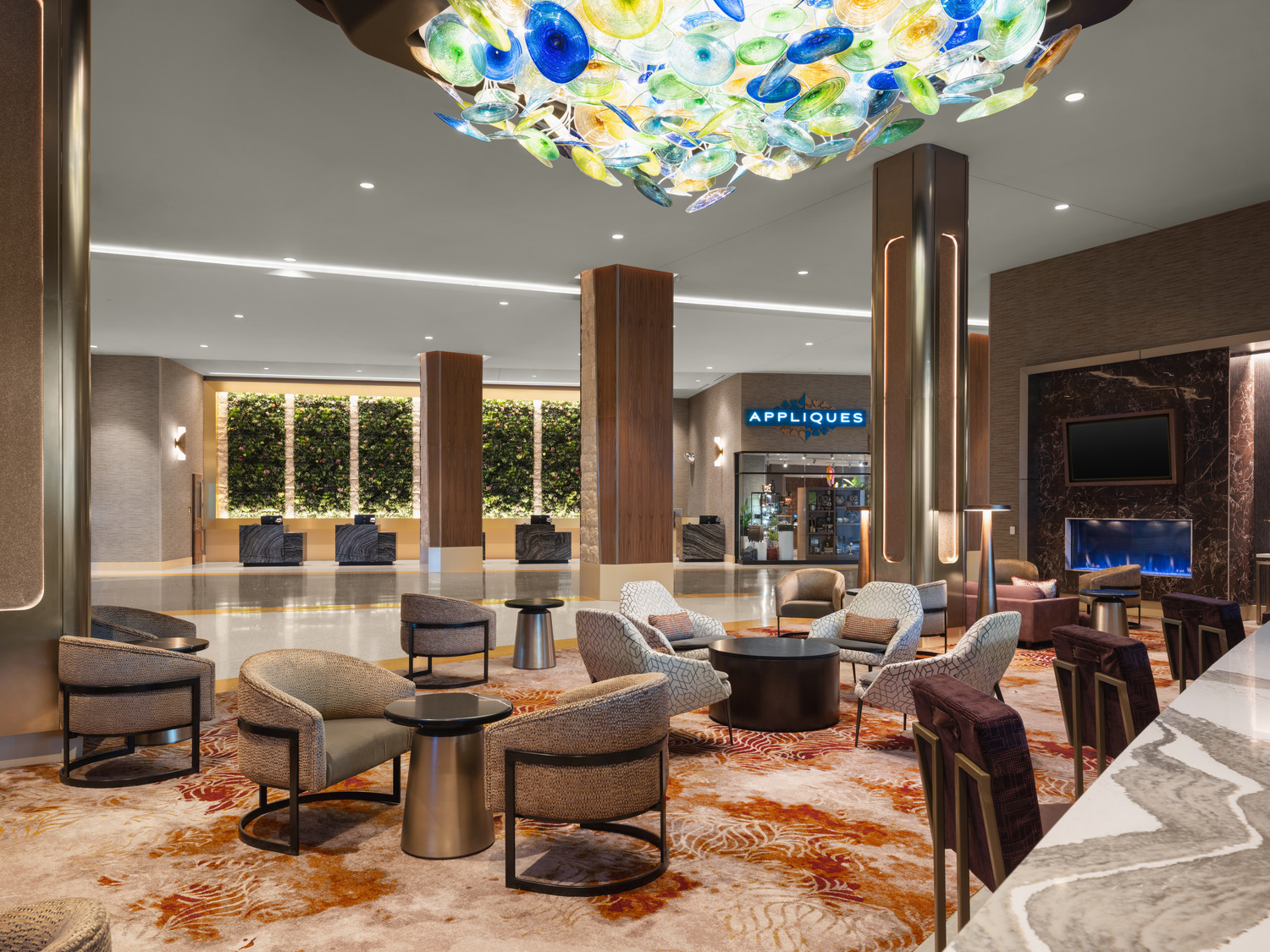 Modern hotel lobby with plush seating, a colorful glass ceiling fixture, vertical plant wall, and a fireplace; restaurant sign 