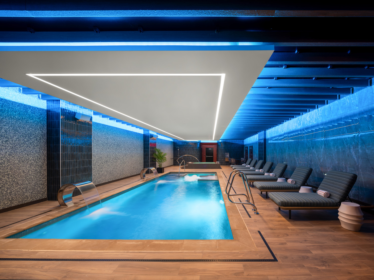 Modern indoor pool area with loungers along one side, contemporary ceiling lights, wooden flooring, and water features at the end of the pool.