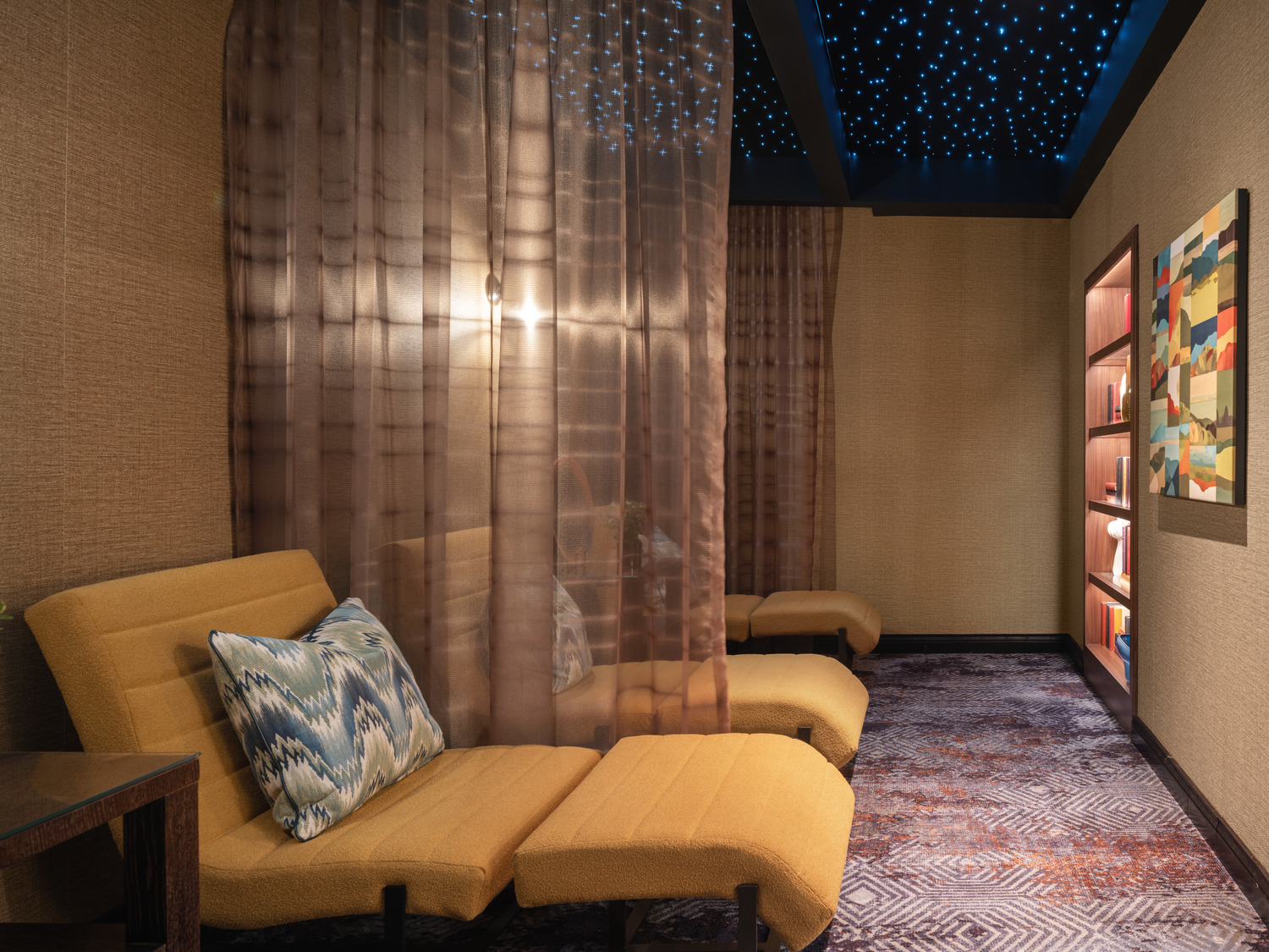 A relaxation lounge with yellow recliners, patterned pillows, sheer curtains, textured walls, a colorful painting, and a ceiling with blue star-like lights.