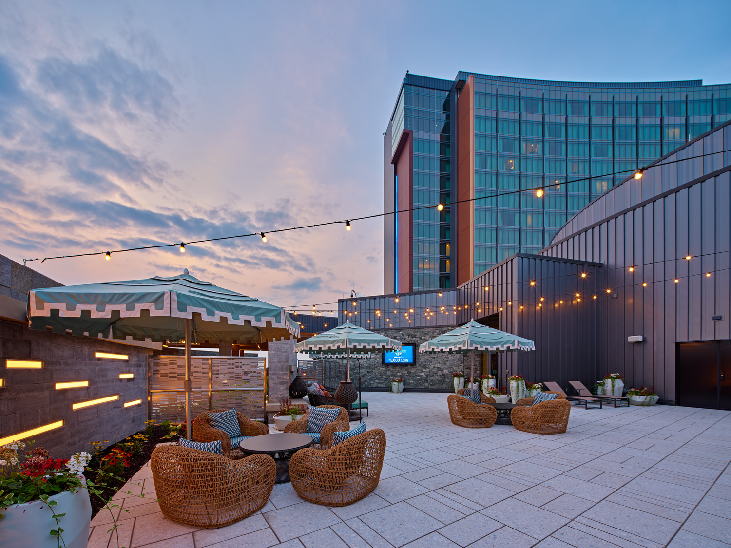 Rooftop patio with wicker seating, striped umbrellas, string lights, potted plants, and a modern glass building in the background at dusk.