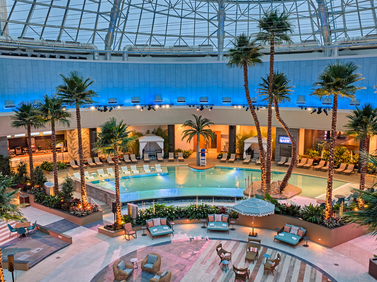Indoor resort pool area with palm trees, lounge chairs, cabanas, and seating areas, enclosed by a glass dome ceiling with blue accent lighting.